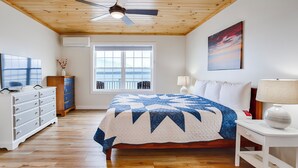 Une chambre avec un grand lit, un ventilateur de plafond, une fenêtre avec vue, une commode, une lampe et un tableau.