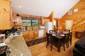 A wooden cabin kitchen with granite countertops, a dining table set for four, and a staircase leading to a loft bedroom.