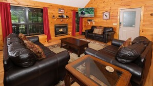 Interior de una cabaña de troncos con sofás de cuero, chimenea, televisor y puerta con panel de vidrio.