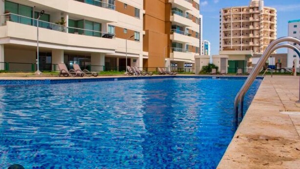 Una piscina con lettini, una zona relax a bordo vasca e un edificio a più piani con balconi sullo sfondo.