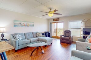 A living room with a sectional sofa, a glass coffee table, a ceiling fan, a TV, and a view of the outdoors through a sliding glass door.
