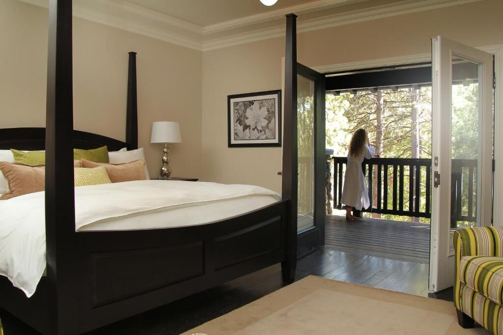 A bedroom with a four-poster bed, a nightstand with a lamp, a framed picture on the wall, and a balcony with a view of trees.