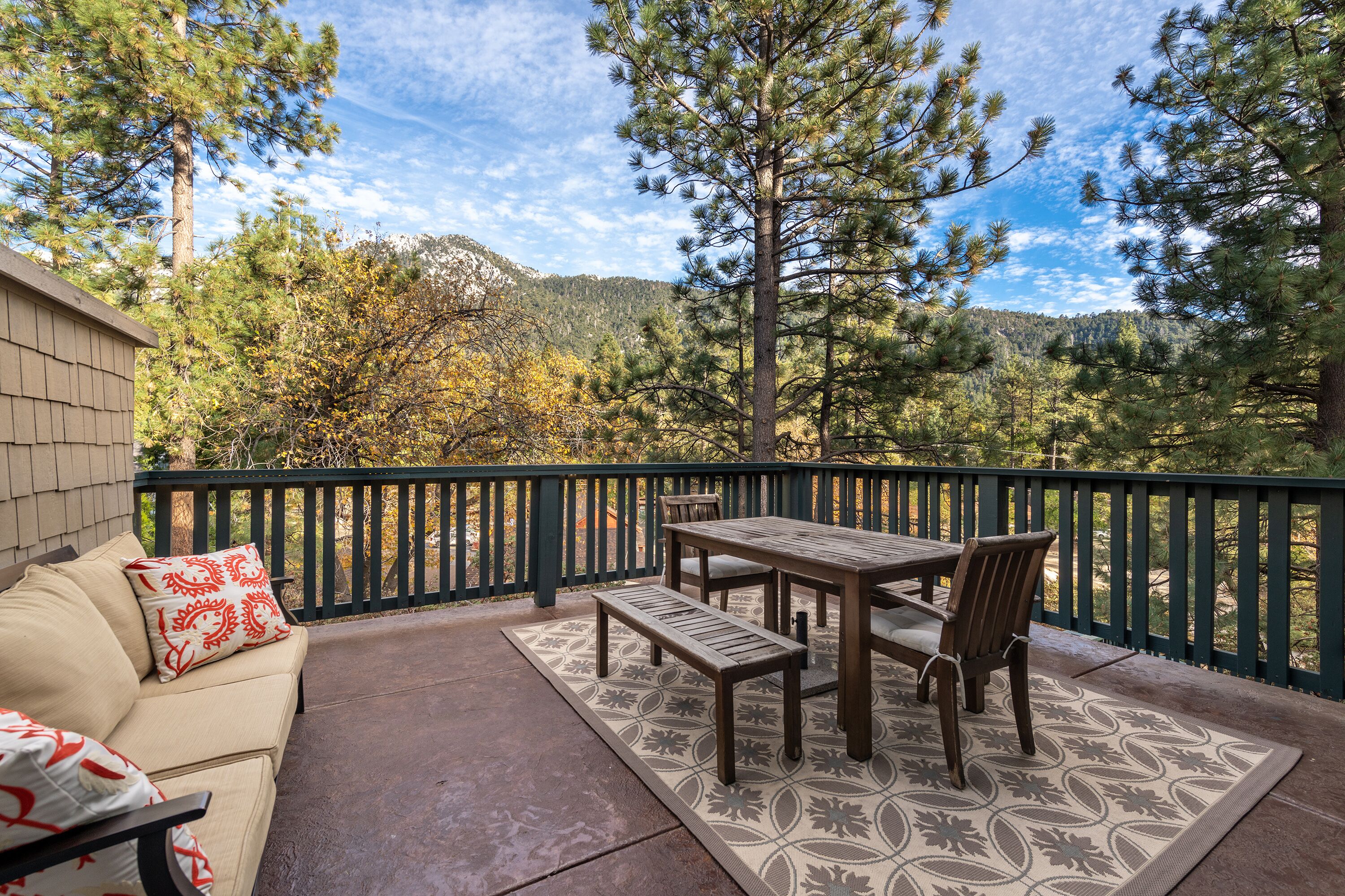 An outdoor patio with a wooden table and benches, surrounded by a green railing, overlooking a forested area with mountains in the background.