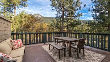 An outdoor patio with a wooden table and benches, surrounded by a green railing, overlooking a forested area with mountains in the background.