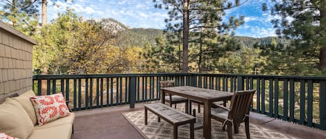 An outdoor patio with a wooden table and benches, surrounded by a green railing, overlooking a forested area with mountains in the background.