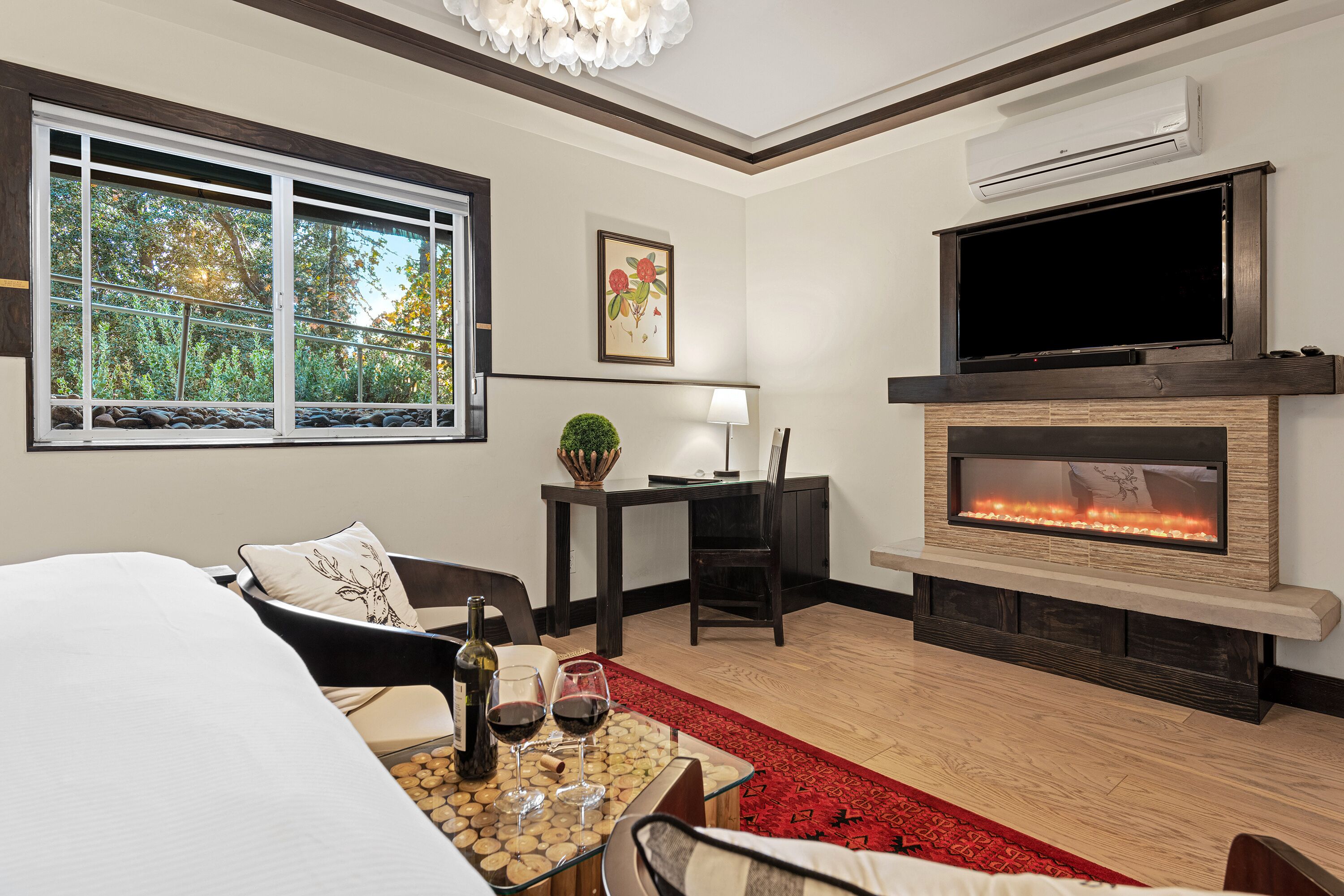 A modern hotel room with a large window, a fireplace, a flat-screen TV, and a dining area with a red rug.