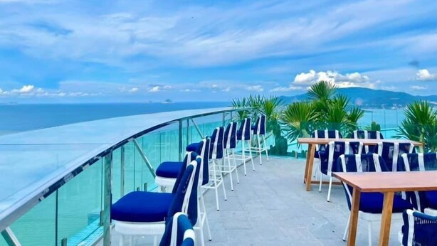 A terrace with blue cushioned chairs and tables, overlooking a body of water and a distant mountain range.