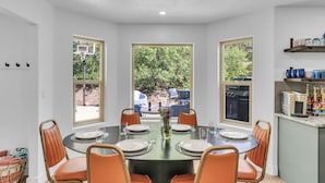 A dining area with a round table, six orange chairs, and a vase of flowers. There are two windows providing views of the outdoors. - Zion Luxe Family Escape: HotTub, Games, Sleeps 14 (Glendale)