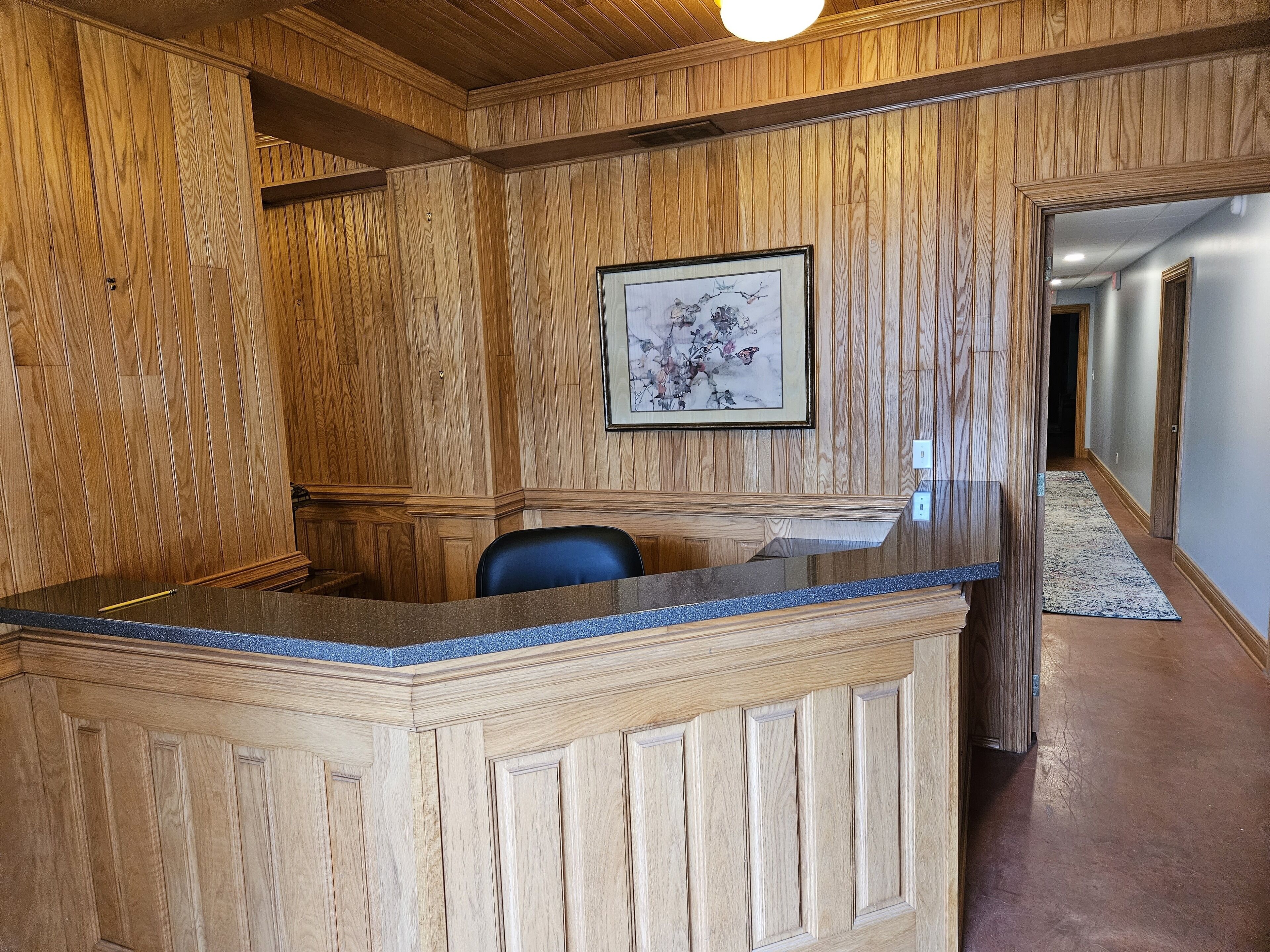 A reception area with a wooden reception desk, a framed artwork on the wall, and a hallway leading to other rooms.
