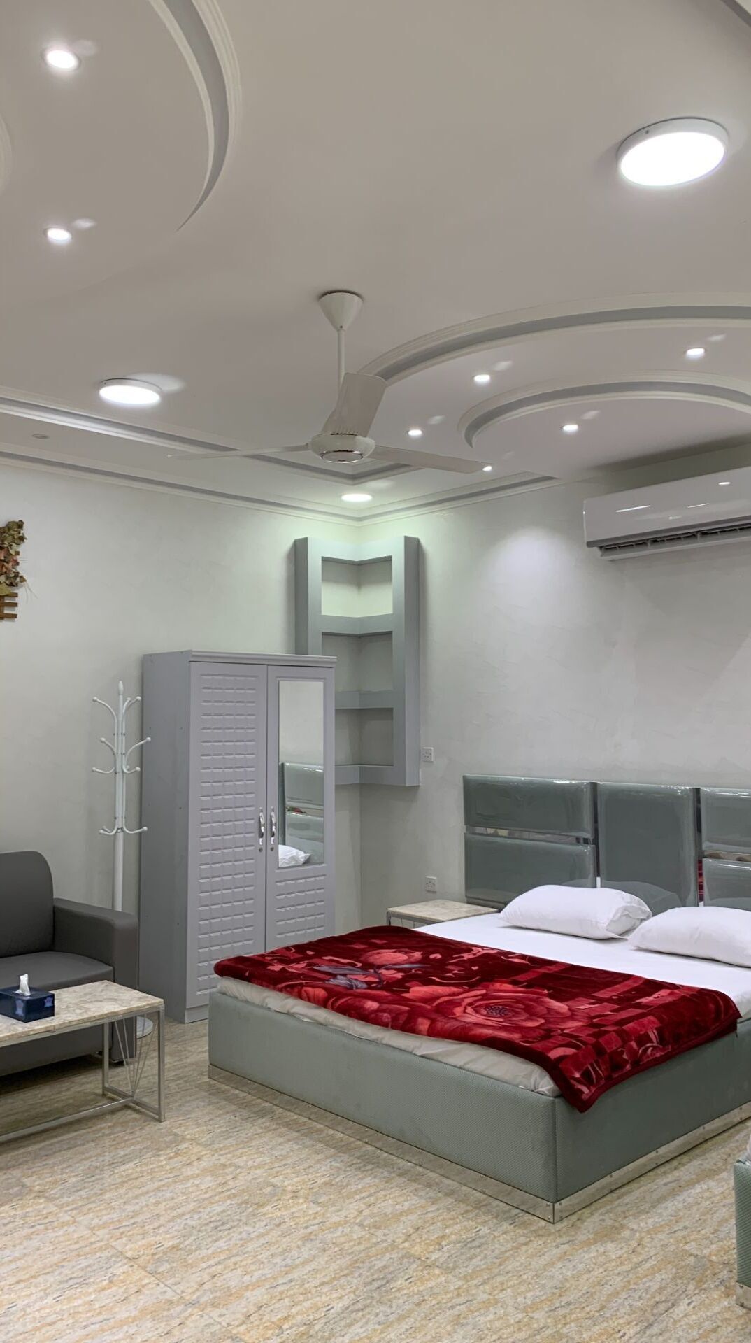 Une chambre d’hôtel moderne avec un lit, une armoire grise, une petite table et un ventilateur de plafond.