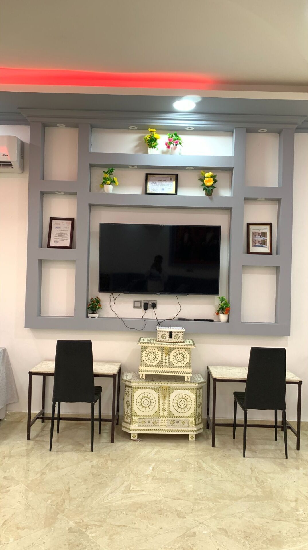 A modern living room with a large flat-screen TV mounted on a grey wall, flanked by two black chairs and a decorative cabinet.