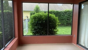 A room with large windows, a ceiling fan, and a view of a green bush and a yellow building. - El Palacio de la Pesca (Lake Placid)