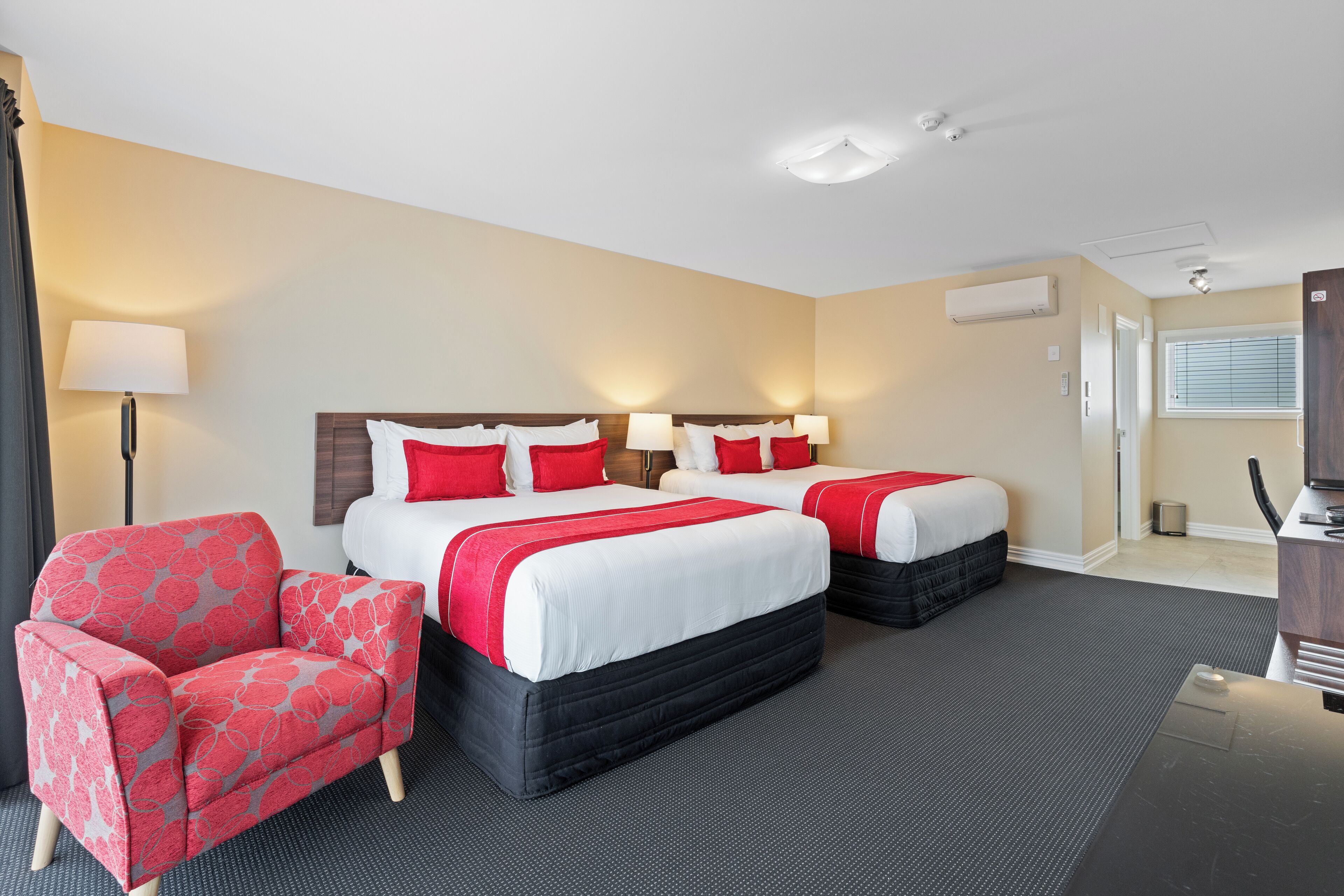 Ein Hotelzimmer mit zwei Betten, einem roten und grauen Sessel, einem Schreibtisch und einer Lampe.
