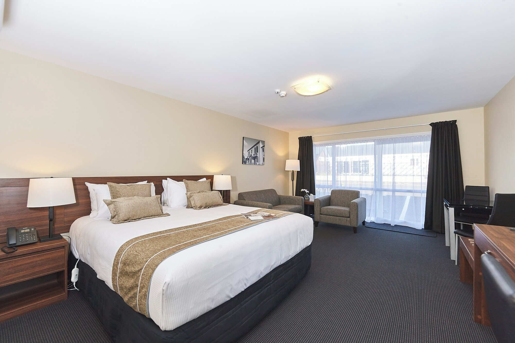 A hotel room with a large bed, two bedside tables, a desk, a chair, a TV, and a window with curtains.