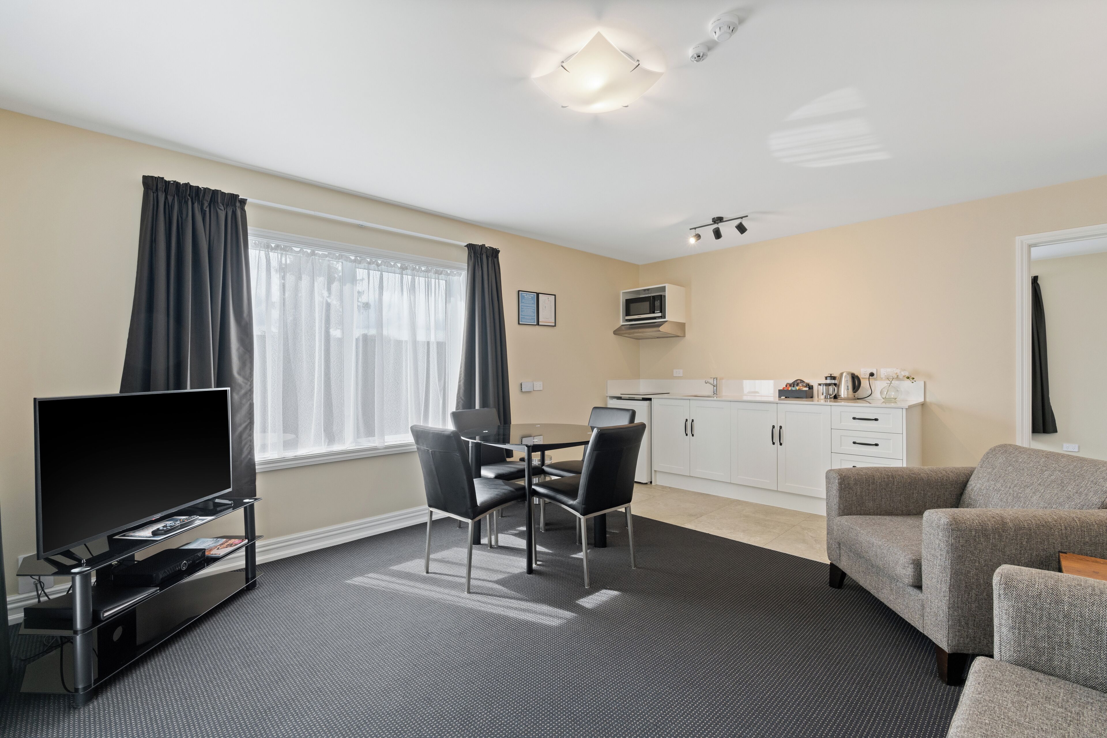 A modern living room with a flat-screen TV, a dining table with four chairs, a gray sofa, and a kitchenette with cabinets and a microwave.