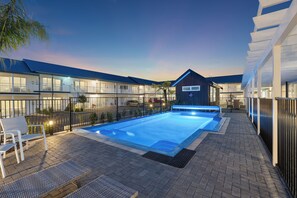 A swimming pool with a lounge area, a covered patio, and a building with multiple windows and balconies.