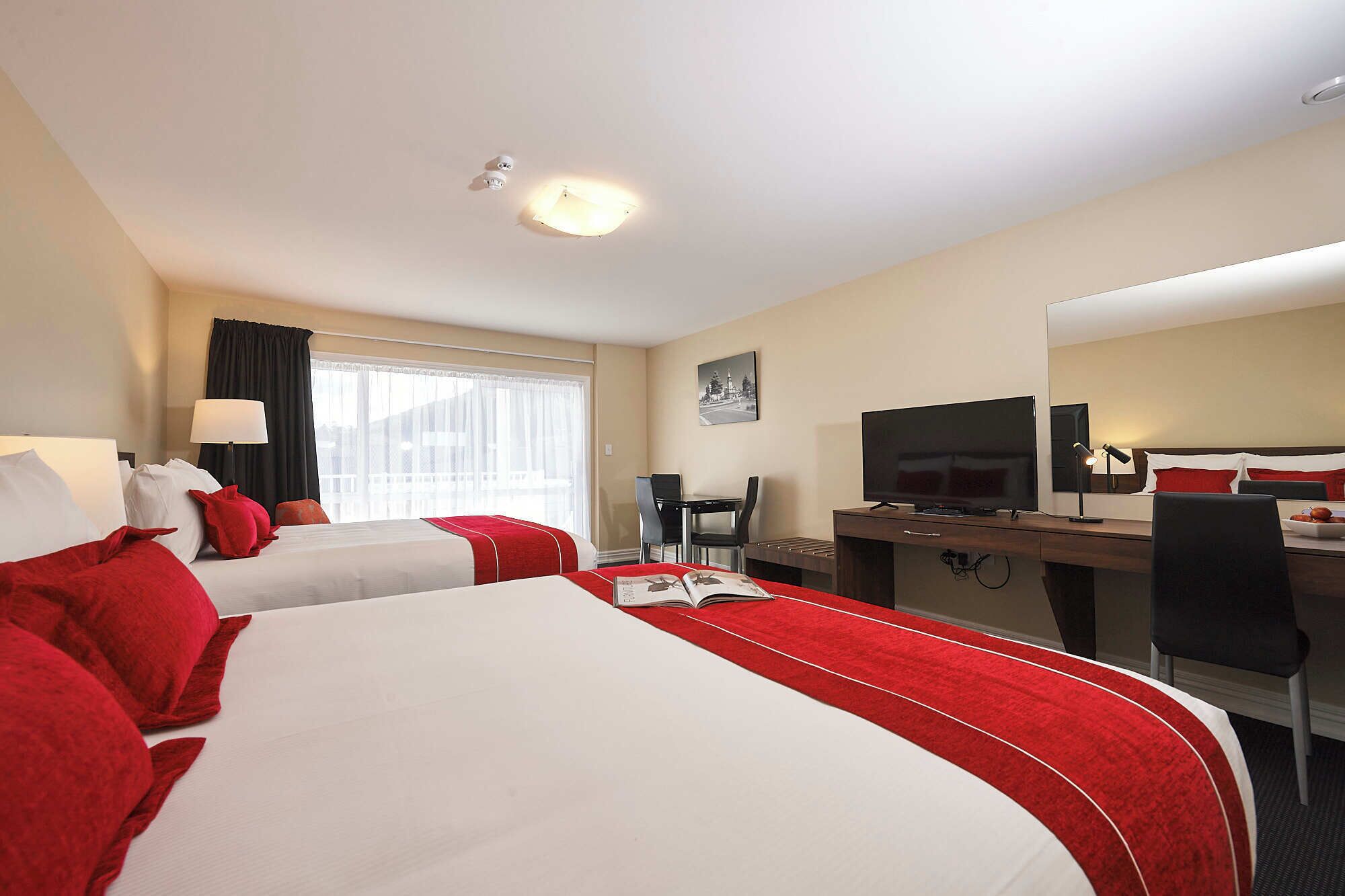 A hotel room with two beds, a desk, a TV, and a dining table.