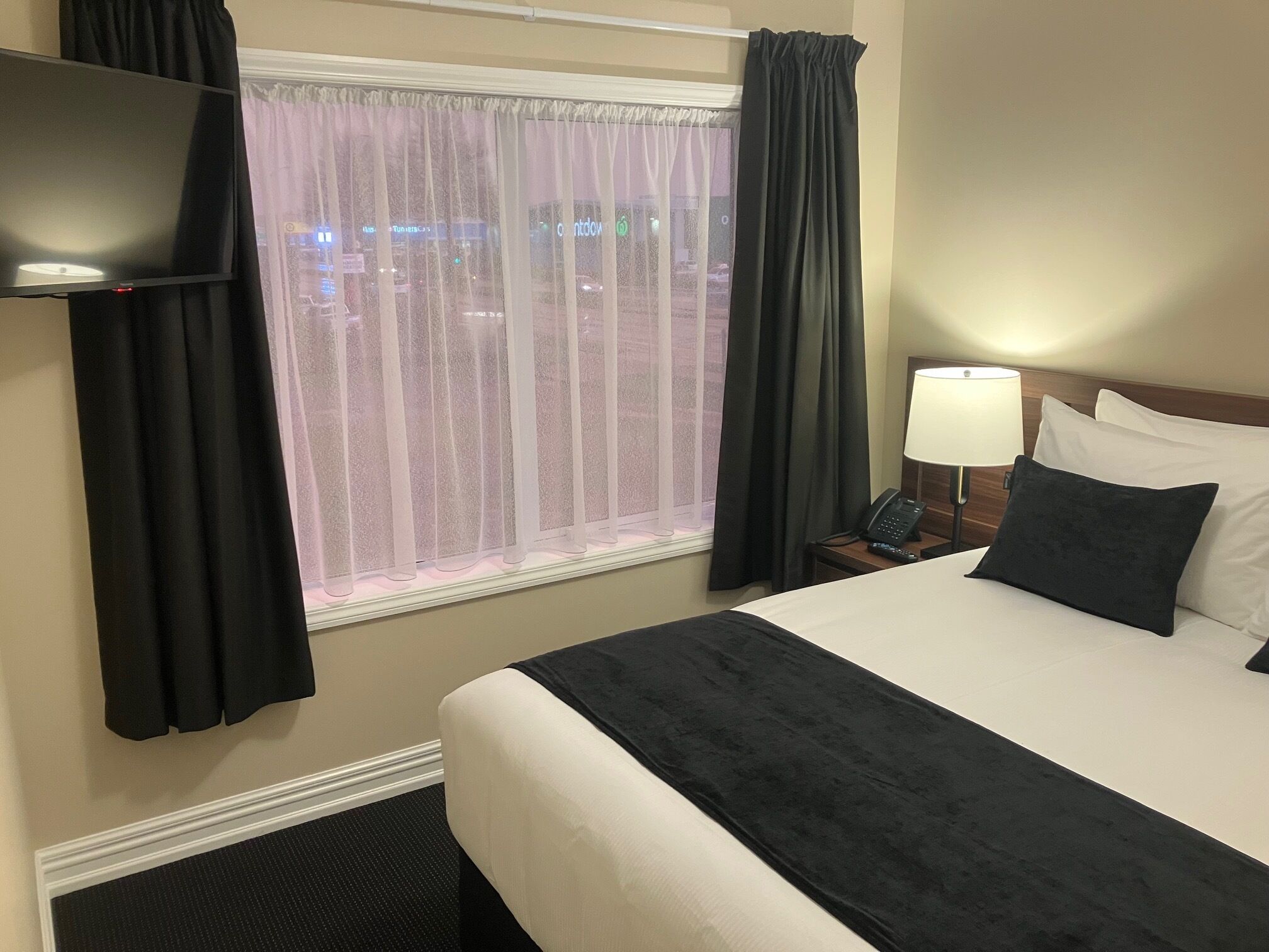 A hotel room with a bed, a TV, a nightstand with a lamp, and a window with curtains.