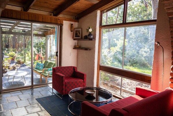 A cozy room with a red armchair, a red sofa, and a glass coffee table. There is a sliding glass door leading to an outdoor patio with a green chair and a table.