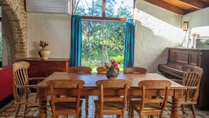 Een eetruimte met een houten tafel en stoelen, een vaas met bloemen en uitzicht op groen door een raam.
