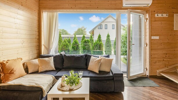 Un salon chaleureux avec un canapé gris foncé, une table basse blanche agrémentée de bougies et d’une plante, et une vue sur une maison et des arbres, le tout visible à travers une grande fenêtre.