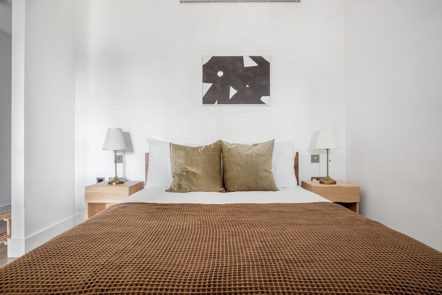 A bedroom with a bed, two nightstands, two lamps, a wall art piece, and a textured brown throw blanket.