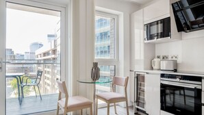 Une cuisine moderne équipée d’un four encastré, d’un four à micro-ondes et d’une machine à café. On y trouve une table en verre avec deux chaises et un vase. La pièce est dotée de grandes fenêtres offrant une vue sur la ville.