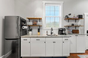 A modern kitchen with white cabinets, a stainless steel refrigerator, a window with blinds, and wooden shelves with kitchen items. - Springside Bourbon Getaway (Georgetown)
