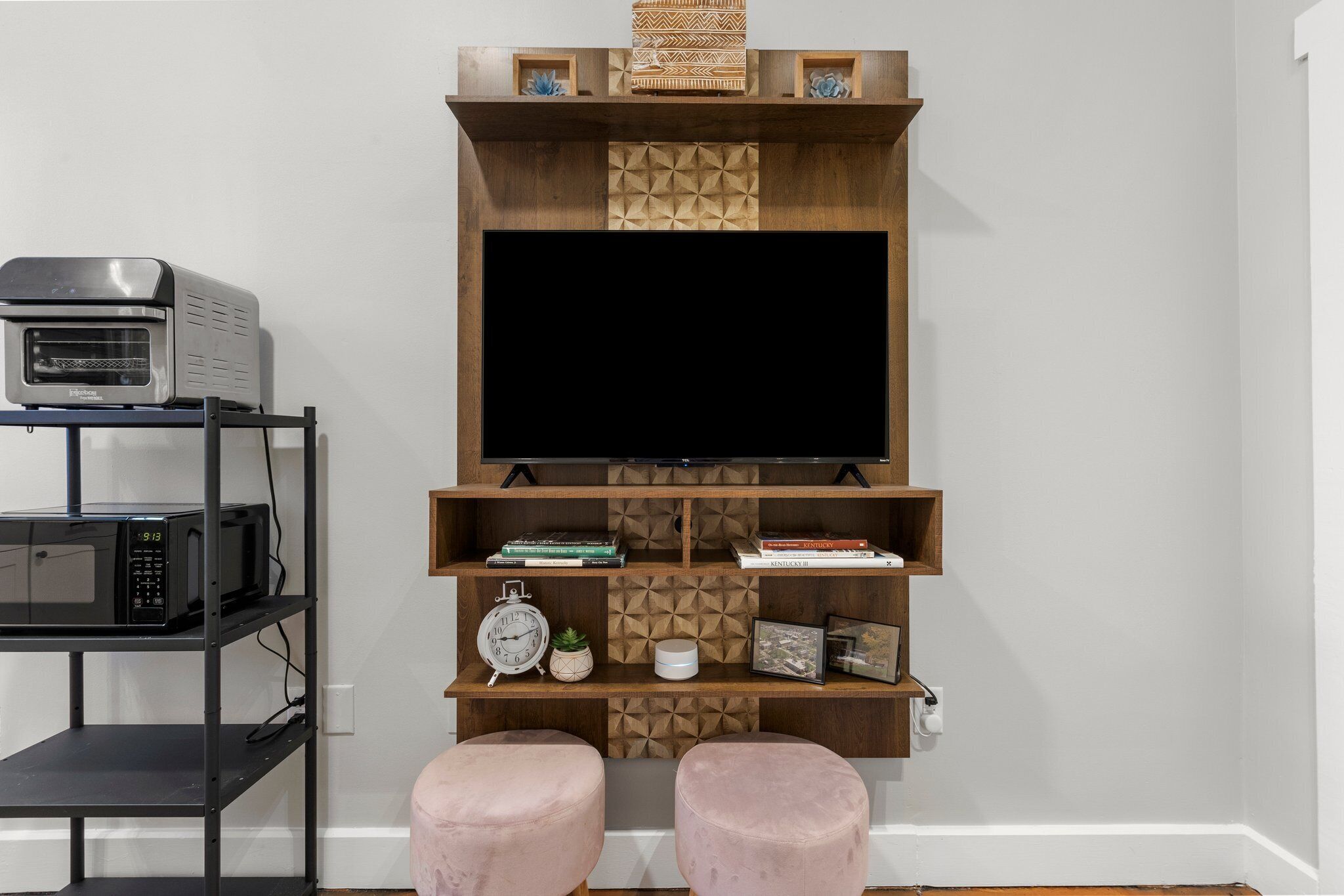 A modern living room with a flat-screen TV mounted on a wooden wall unit, a microwave on a shelf, and two pink ottomans.