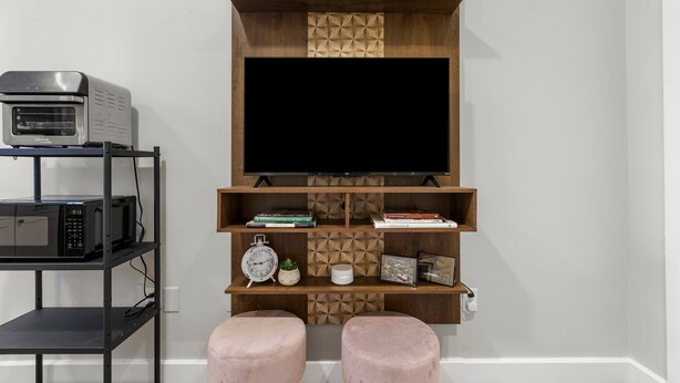 Un salon moderne avec un téléviseur à écran plat fixé à un meuble mural en bois, un four à micro-ondes posé sur une étagère et deux poufs roses.