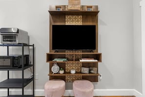 A modern living room with a flat-screen TV mounted on a wooden wall unit, a microwave on a shelf, and two pink ottomans. - Springside Bourbon Getaway (Georgetown)