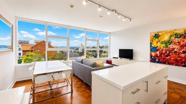 A modern living room with a dining area, large windows, a flat-screen TV, and a colorful abstract painting.