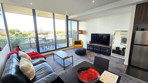 Una sala de estar moderna con un sofá de cuero, una mesa de comedor de vidrio, un televisor de pantalla plana grande y una zona de cocina con refrigerador.