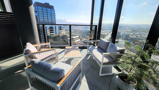 A high-rise balcony with modern furniture, including a sofa and a table, overlooking a cityscape with buildings and a clear sky.