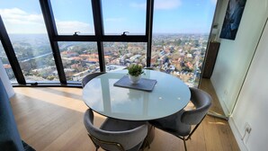 A modern room with a round glass table, two grey upholstered chairs, a potted plant, and a view of a cityscape through large windows.