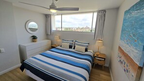 A bedroom with a large bed, a ceiling fan, a window with curtains, a bedside table with a lamp, a small round mirror, and a painting on the wall. - Stunning Manly Beach views - 906 Manly National (Manly)