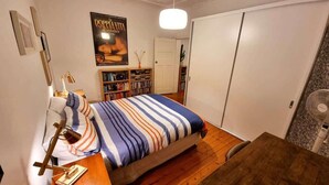 Een slaapkamer met een bed, nachtkastje, lamp, boekenkast en een poster aan de muur.