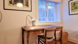 Een kamer met een houten bureau, een stoel, een raam met witte luiken en een lijst met een afbeelding aan de muur.