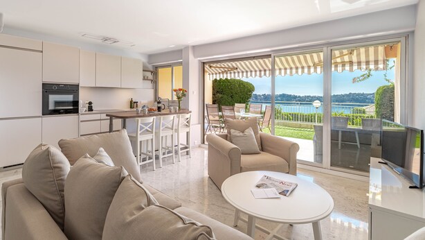 A modern living room with a sofa, a coffee table, and a TV. There is a kitchen area with cabinets and a dining table. The room has large windows with a view of a lake and greenery. - Flora Sur Mer 2 bedroom apartment (Villefranche-sur-Mer)