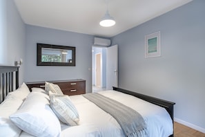 A bedroom with a bed, bedside table, dresser, mirror, and an air conditioning unit.