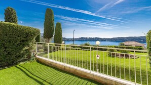 A well-maintained lawn with a white metal fence, a clear blue sky, and a distant view of a lake and buildings. - Flora Sur Mer 2 bedroom apartment (Villefranche-sur-Mer)