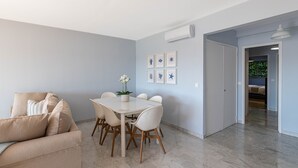 A modern living room with a beige sofa, a white dining table with six chairs, and a potted plant. There are framed pictures on the wall, and an air conditioning unit is mounted on the wall. - Flora Sur Mer 2 bedroom apartment (Villefranche-sur-Mer)