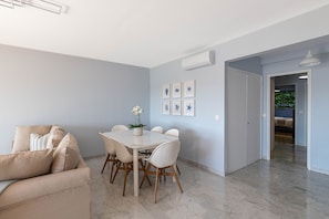 Una sala de estar moderna con un sofá beige, una mesa de comedor blanca con seis sillas y una planta en maceta. Hay cuadros enmarcados en la pared, y un equipo de aire acondicionado está instalado allí.