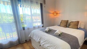 Une chambre à coucher avec un lit, une fenêtre avec des rideaux, un luminaire suspendu et une vue sur la verdure à l’extérieur.
