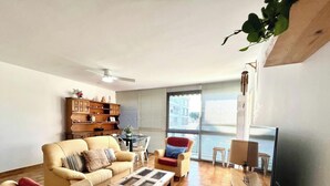 Un salon comprenant un canapé, une table basse, une bibliothèque, un fauteuil et une fenêtre avec des stores.