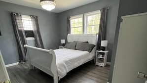 Ein Schlafzimmer mit Bett, Nachttisch, Lampe, Deckenventilator und Fenster mit Vorhängen.
