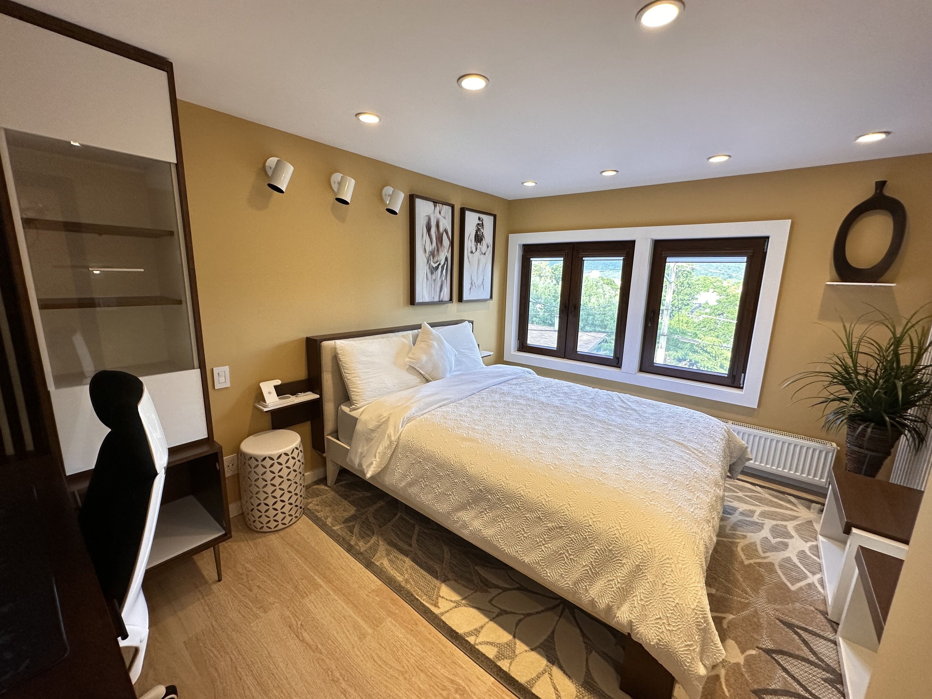 A bedroom with a large bed, a nightstand, a lamp, a potted plant, and a window with a view of greenery.