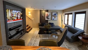 Ein modernes Wohnzimmer mit Fernseher, der Nachrichten zeigt, einem grauen Sofa, einem Couchtisch und einer Treppe, die zu einer anderen Ebene führt.