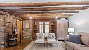 Un salon chaleureux avec des poutres apparentes, un mur en briques et une cheminée en pierre. On y trouve deux fauteuils ornés de coussins à motifs floraux, un canapé et un meuble en bois.