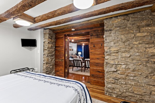 A bedroom with a bed, wooden beams, stone walls, a television, and a doorway leading to a living area with a sofa and chairs. - Springside Equine Getaway (Georgetown)